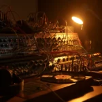 Analog modular synthesizer rig in a dimly lit home studio with patch cables, a vinyl record and headphones beside a small mixing console.