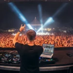 View from behind a DJ on stage facing a massive festival crowd with dramatic blue and white lights.