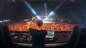 View from behind a DJ on stage facing a massive festival crowd with dramatic blue and white lights.