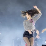 Olivia Rodrigo performing live, mid-motion with her hair flying, in front of a smoke-filled arena stage