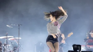 Olivia Rodrigo performing live, mid-motion with her hair flying, in front of a smoke-filled arena stage