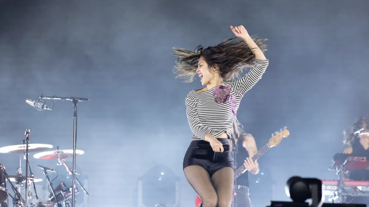 Olivia Rodrigo performing live, mid-motion with her hair flying, in front of a smoke-filled arena stage