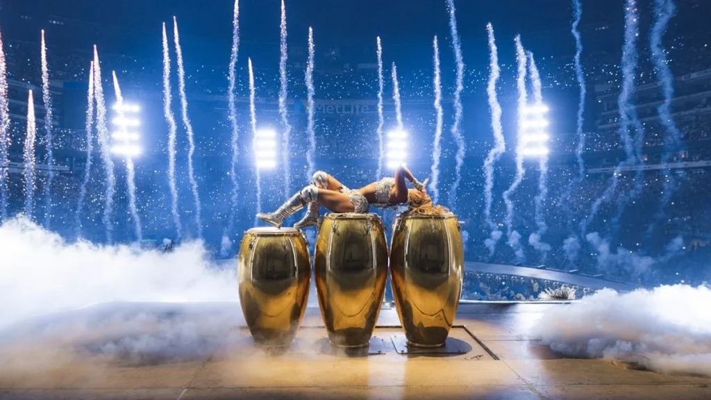Karol G lies across gold conga drums under pyrotechnics at MetLife Stadium